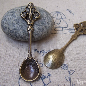 Può includere: Due cucchiaini in bronzo di stile antico con manici decorati. I cucchiaini sono piccoli e hanno un design decorativo.