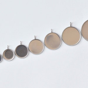 May include: A collection of round pendant trays in various sizes, arranged diagonally. Each pendant has a silver-colored bezel and a loop for attaching a chain. The interior of the pendants varies in color, from dark grey to light beige.