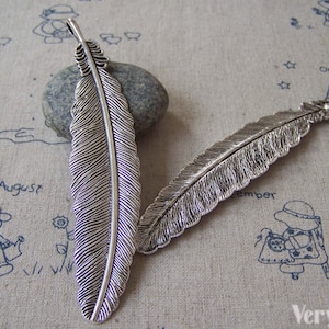 May include: Two silver feather charms with detailed textures. The charms are about 2 inches long and have a loop at the top for attaching to jewelry.