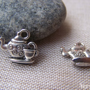 May include: Two silver charm pendants shaped like teapots with a cup and saucer. The teapots have a swirl design.