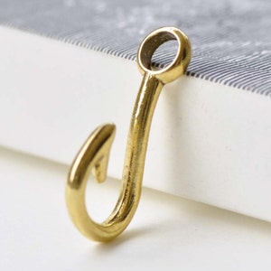 Pode incluir: Um amuleto de anzol de pesca em metal dourado. O anzol é curvo e tem um pequeno laço na parte superior.