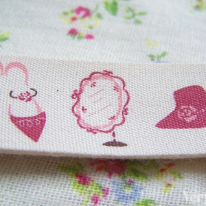 Könnte beinhalten: Weißer Stoff mit rosafarbenem Blumendruck und einem rosafarbenen Band mit drei rosafarbenen Bildern: ein Paar Schuhe, ein Spiegel und ein Hut.