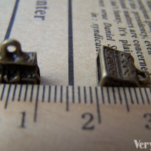 May include: Two antique gold-toned metal charms, one shaped like a stack of books and the other like a single book, are shown on a ruler. The ruler measures in centimeters and shows the charms are less than 1 centimeter in size.