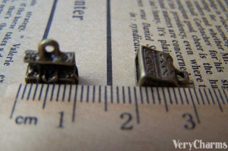 May include: Two antique gold-toned metal charms, one shaped like a stack of books and the other like a single book, are shown on a ruler. The ruler measures in centimeters and shows the charms are less than 1 centimeter in size.