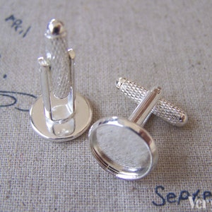 May include: Two silver-toned cufflinks with a textured design on the bar and post. The cufflinks have a round base and a smooth, polished finish. The background is a light-colored fabric.