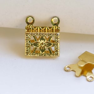 Op de afbeelding: Gouden metalen sluiting met een decoratief bloemmotief. De sluiting heeft aan elke kant twee lussen om sieraden aan te bevestigen.