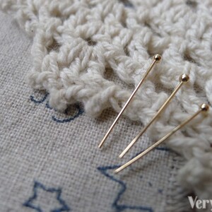 May include: Three gold-colored, straight, thin, metal pins with round heads. The pins are lying on a white crocheted doily.