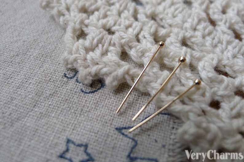 May include: Three gold-colored, straight, thin, metal pins with round heads. The pins are lying on a white crocheted doily.