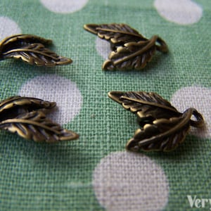 May include: Four antique gold-toned leaf charms with a twisted wire design. The charms are on a green fabric background with white polka dots.