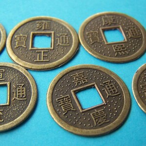 K&ouml;nnte beinhalten: Sechs antike chinesische M&uuml;nzen aus Messing mit einem quadratischen Loch in der Mitte. Die M&uuml;nzen sind mit chinesischen Schriftzeichen verziert. Die M&uuml;nzen liegen auf einem blauen Hintergrund.