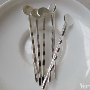 Pu&ograve; includere: Sei fermagli per capelli argentati con decorazioni rotonde in metallo argentato nella parte superiore. I fermagli sono disposti in una pila su un piatto bianco.