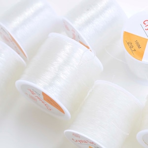 Puede incluir: Seis carretes de hilo elástico transparente, cada uno etiquetado con "Crystal Tec Korea 100M 0.7".