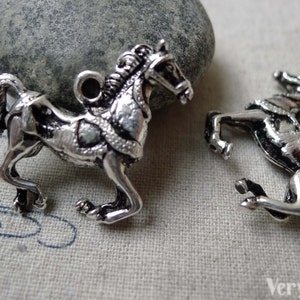 May include: Two silver-colored horse charms. One horse is in a trotting pose, and the other is rearing up. Both charms have detailed manes, harnesses, and a loop for attaching to a necklace or bracelet. The charms are on a light-colored fabric.