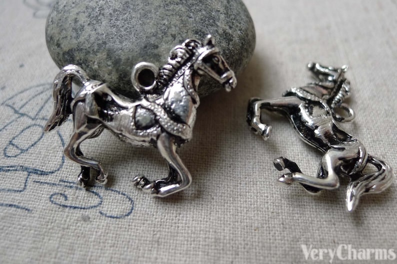 May include: Two silver-colored horse charms. One horse is in a trotting pose, and the other is rearing up. Both charms have detailed manes, harnesses, and a loop for attaching to a necklace or bracelet. The charms are on a light-colored fabric.