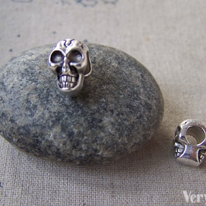 May include: Two silver skull beads, one with a hole through the back and the other with a hole through the top. The beads are on a grey stone surface.