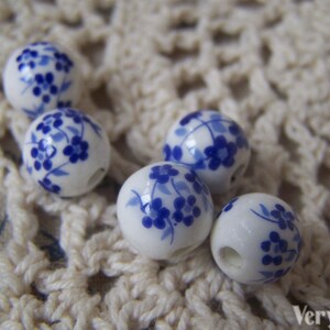 Puede incluir: Seis cuentas de cer&aacute;mica blanca con dise&ntilde;os florales azules. Las cuentas son redondas y tienen un agujero en el centro. Est&aacute;n dispuestas sobre una superficie de crochet blanco.