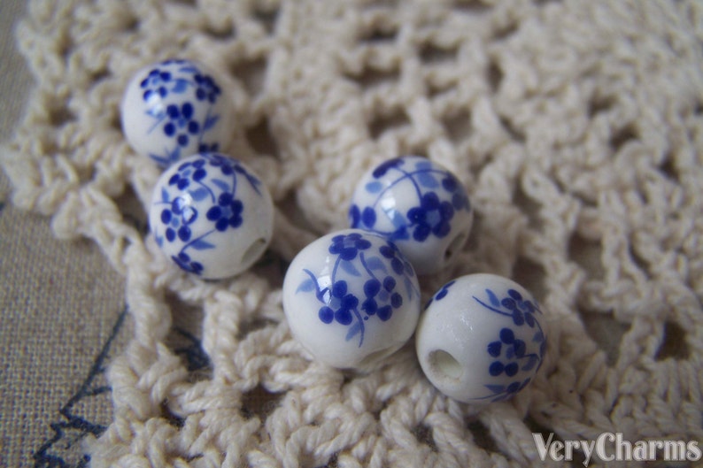 Puede incluir: Seis cuentas de cer&aacute;mica blanca con dise&ntilde;os florales azules. Las cuentas son redondas y tienen un agujero en el centro. Est&aacute;n dispuestas sobre una superficie de crochet blanco.