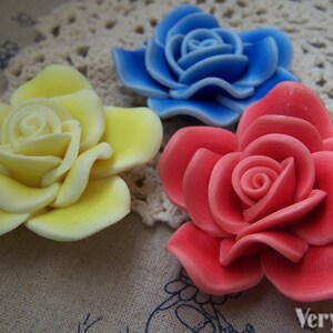 Peut inclure: Trois roses en argile polym&egrave;re faites &agrave; la main en jaune, bleu et rouge. Les roses sont pr&eacute;sent&eacute;es sur une dentelle blanche au crochet.