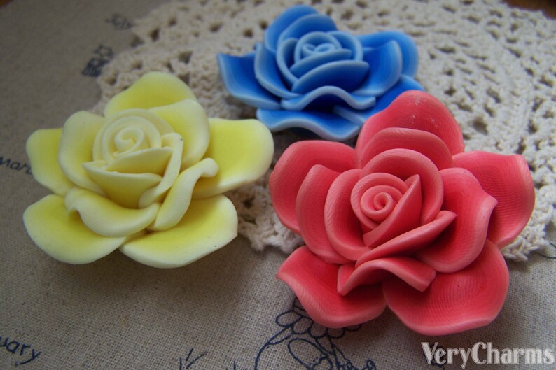 Peut inclure: Trois roses en argile polym&egrave;re faites &agrave; la main en jaune, bleu et rouge. Les roses sont pr&eacute;sent&eacute;es sur une dentelle blanche au crochet.