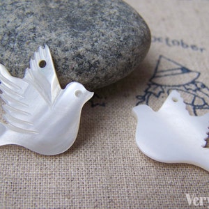 May include: Two white mother of pearl dove charms. The dove on the left has detailed wings and a hole for hanging. The dove on the right is a simpler design.