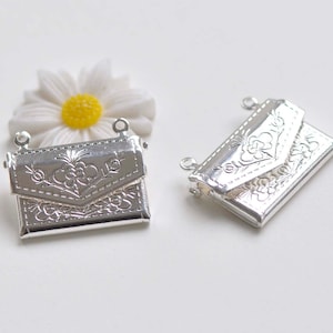 Könnte beinhalten: Zwei silberfarbene Metall-Anhänger in Form einer Handtasche mit Blumenmustern. Die Anhänger haben oben zwei Schlaufen zum Anbringen an einer Halskette oder einem Armband.