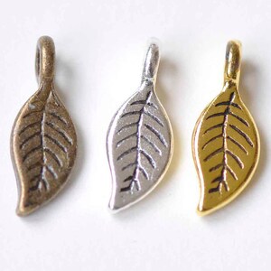 Puede incluir: Tres abalorios de metal en forma de hoja en oro, plata y bronce. Cada abalorio tiene un diseño de hoja detallado.