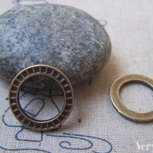 May include: Two antique brass-coloured metal rings. One ring has a detailed, patterned design around the outer edge, while the other is a simple, smooth band. A grey stone rests in the background.