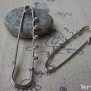 Puede incluir: Dos imperdibles plateados con bucles decorativos en el imperdible. Los imperdibles están sobre un fondo de tela.