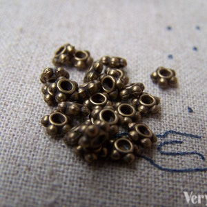 May include: A close-up of a pile of small, gold-colored metal beads. The beads are round with a small hole in the center and have a decorative design around the edge.