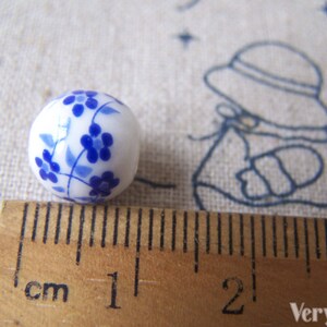 Puede incluir: Una cuenta de cer&aacute;mica blanca con un dise&ntilde;o floral azul. La cuenta mide aproximadamente 1 cm de di&aacute;metro.
