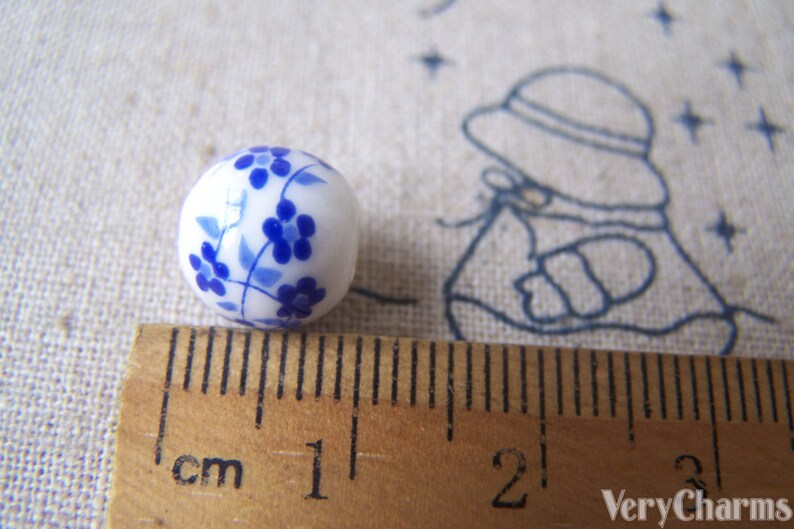 Puede incluir: Una cuenta de cer&aacute;mica blanca con un dise&ntilde;o floral azul. La cuenta mide aproximadamente 1 cm de di&aacute;metro.