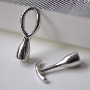 May include: Two silver metal bottle stoppers with unique designs. One stopper has a loop at the top and a rounded bottom. The other stopper has a hook-like shape at the top and a rounded bottom.