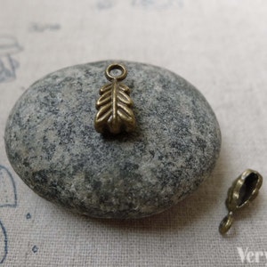 May include: A bronze leaf-shaped charm with a small loop at the top. The charm is about 1 inch long and is sitting on a gray rock.
