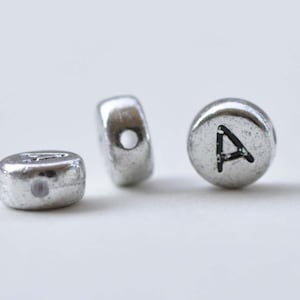 Könnte beinhalten: Drei silberne Metallperlen, wobei eine Perle mit dem Buchstaben 'A' graviert ist. Die Perlen haben ein Loch in der Mitte zum Aufziehen.