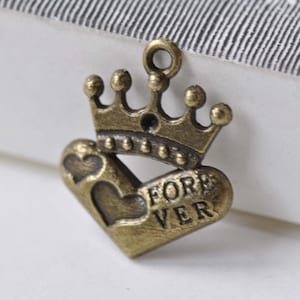 Pode incluir: Um berloque de cor bronze com uma coroa sobre um coração. O coração tem um pequeno recorte em forma de coração e a palavra "FOREVER" gravada. O berloque tem uma argola para prender em joias.