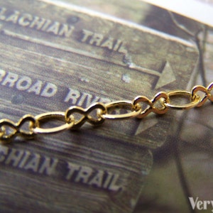 May include: A gold chain necklace with a delicate infinity link design. The chain is lying on a wooden surface with text that reads "Appalachian Trail" and "South Broad River".