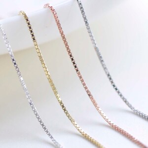 May include: Four silver chains in different colours: silver, gold, rose gold, and a darker silver. The chains are laid out on a white surface.