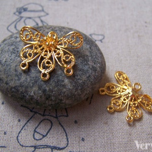 May include: Two gold filigree butterfly charms with three jump rings on each. The charms are on a gray stone surface.