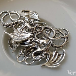 May include: Silver-toned dragon charm with intricate details, featuring a curled tail and outstretched wings. The charm has two jump rings for attaching to jewellery or other crafts.