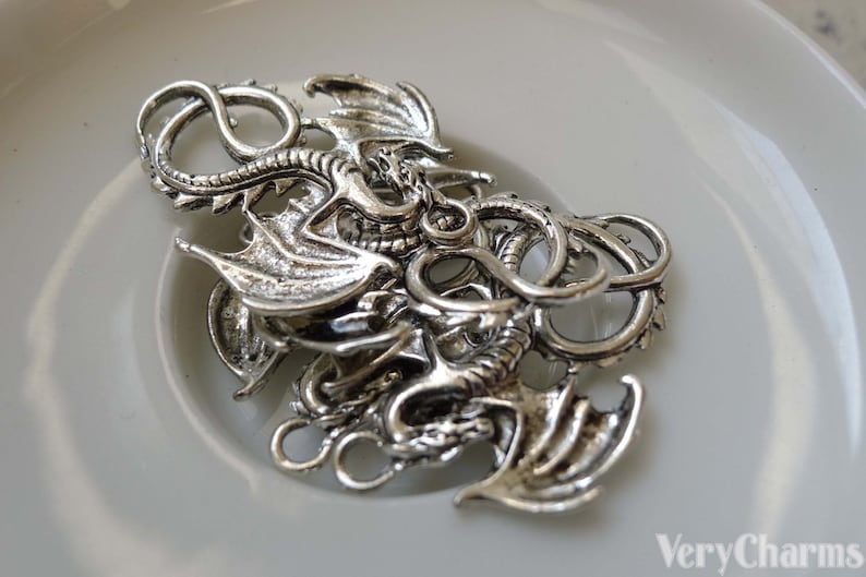 May include: Silver-toned dragon charm with intricate details, featuring a curled tail and outstretched wings. The charm has two jump rings for attaching to jewellery or other crafts.