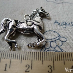 May include: A silver-colored horse charm with intricate details, including a braided mane and a saddle. The horse is in a trotting pose, and a ruler shows it is approximately 1 inch long. The word "August" is visible in the background.