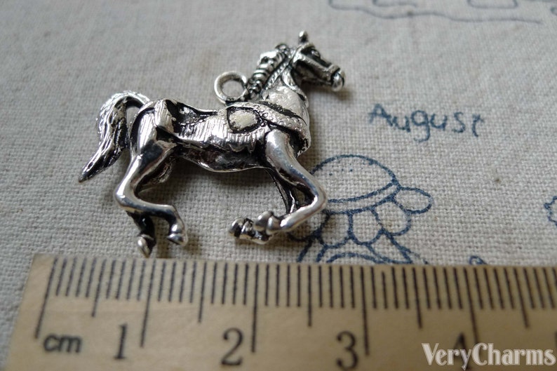 May include: A silver-colored horse charm with intricate details, including a braided mane and a saddle. The horse is in a trotting pose, and a ruler shows it is approximately 1 inch long. The word "August" is visible in the background.