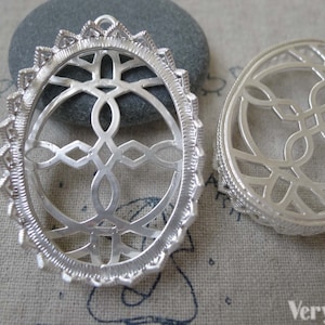 Könnte beinhalten: Zwei silberfarbene Anhänger mit aufwendigen filigranen Designs. Einer ist oval mit dekorativem Rand, der andere rund. Die Anhänger zeigen geometrische und keltische Knotenmuster, geeignet für Schmuckherstellung oder Bastelarbeiten.