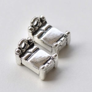 May include: Two silver-toned metal bed charms with a detailed design. The charms are shaped like a bed with a headboard, footboard, and mattress.