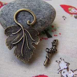 May include: A pair of antique bronze metal charms, one shaped like a grapevine leaf and the other like a bunch of grapes. The charms are on a white fabric background with a pattern of red and white figures.