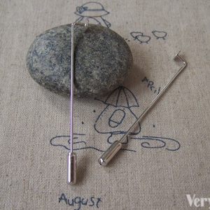 May include: Two silver metal hat pins with rounded ends. The pins are lying on a beige fabric surface with a drawing of a person, an umbrella, and birds.