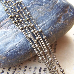 May include: A close-up of a silver chain with small beads. The chain is lying on a gray rock.