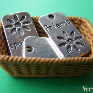 May include: Three silver charm pendants in a wicker basket. The charms feature a flower design, the word "Friends", and the word "Sparkles".