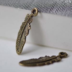Puede incluir: Dos abalorios de plumas de color dorado antiguo con un bucle en la parte superior para sujetar a joyas o manualidades.
