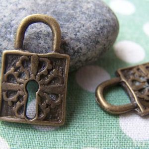 May include: Two bronze-colored metal lock charms with intricate floral designs. One charm is upright, the other is lying down. The charms have a square shape with a keyhole and a curved handle. The background is a green and white polka dot pattern.